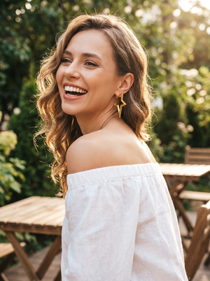 A woman with wavy brown hair smiles outdoors while wearing a white off-the-shoulder top and large gold geometric drop earrings. Wooden patio tables and green foliage are blurred in the background.